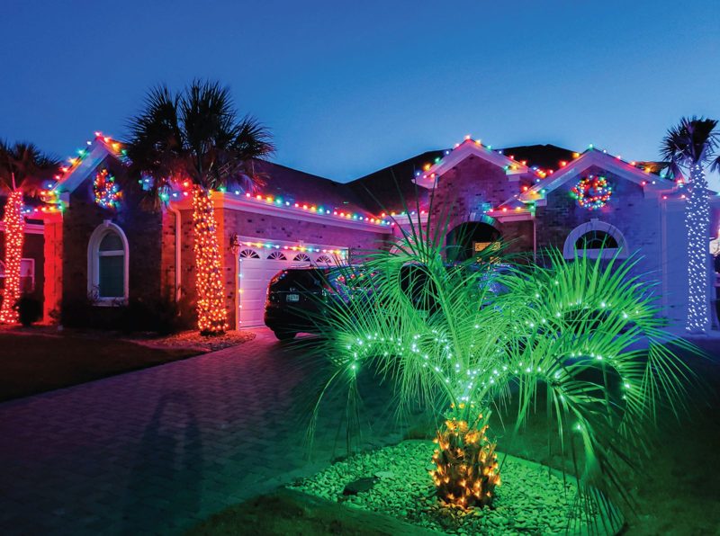 Brick home with warm white roofline lights, a lit palm tree and red-and-green accents