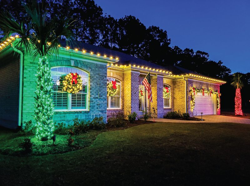 Brick home with warm white roofline lights, lit wreaths on the windows and a wrapped palm