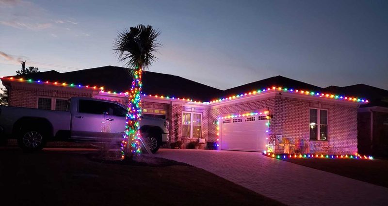 Home with multicolor roofline lights and a lit palm tree at dusk