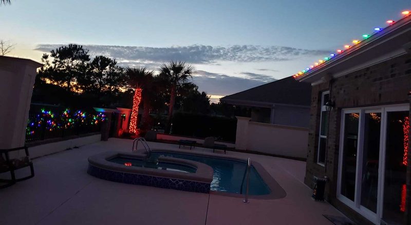 Warm white roofline lights on a home with a lit pool area and a red-wrapped palm