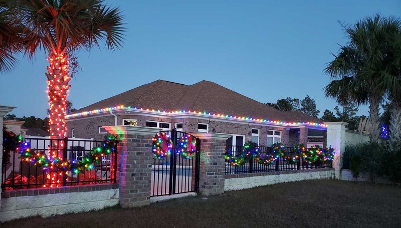 Brick home with multicolor roofline lights and lit wreaths on the fence