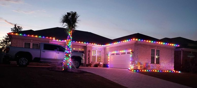 Brick home with multicolor roofline lights and a wrapped palm at dusk