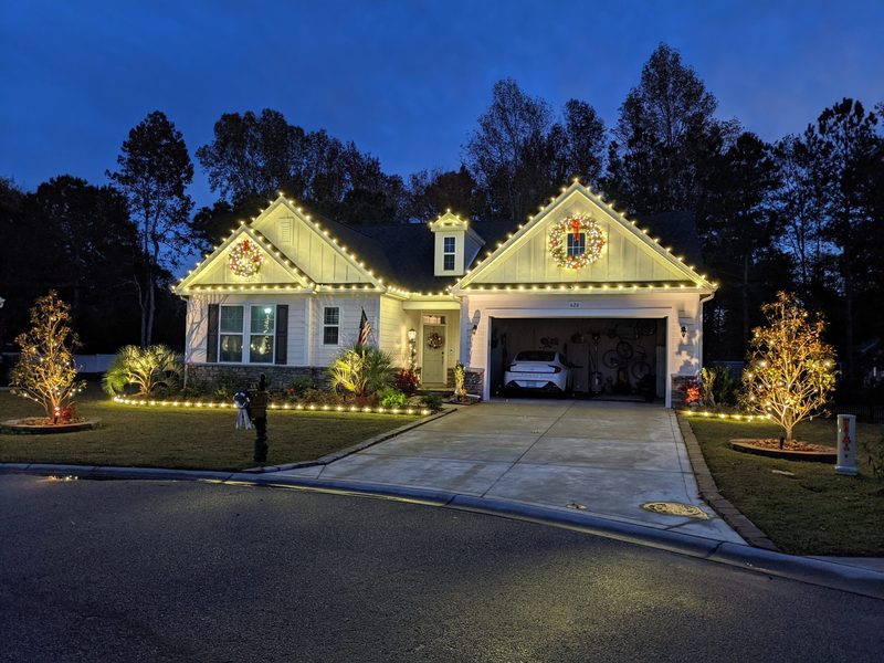 Two-story home with warm white roofline lights and a lit wreath over the garage