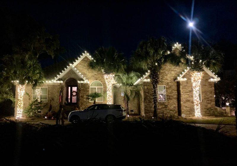 Stucco home with warm white roofline lights and wrapped palm trunks